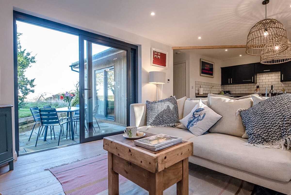 The open plan living area features a spacious sofa adorned with decorative pillows, positioned to face sliding doors leading to a private patio. A wooden coffee table holds magazines, while a dining set with blue chairs is visible outside, nestled against a serene backdrop.