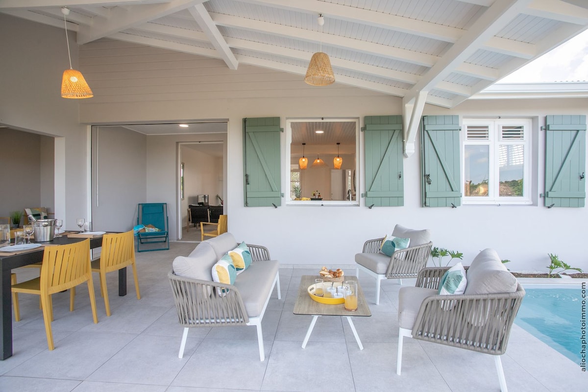 A spacious outdoor seating area is highlighted by comfortable chairs and a coffee table, set under a sloped ceiling with wooden beams. Light green shutters frame the windows, which connect to an open interior. A dining space is visible, alongside a peaceful garden backdrop.