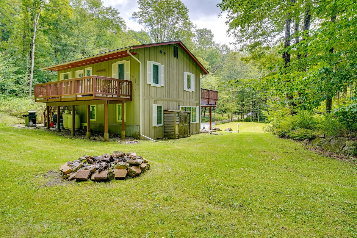 A two-story vacation rental is nestled among trees, featuring a spacious backyard and a fire pit area with stone seating. The building's green exterior is complemented by multiple windows and a large deck above, offering outdoor seating with a natural view.