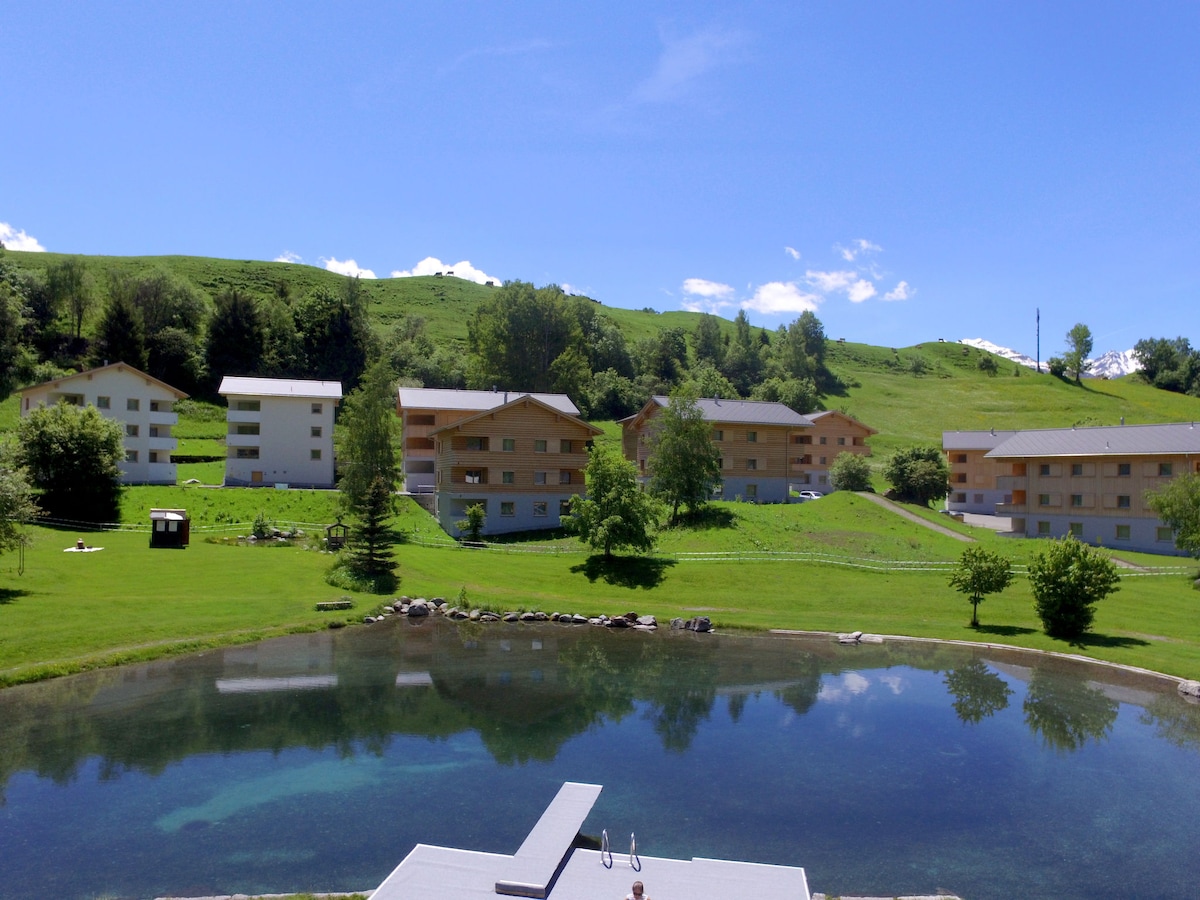 A serene landscape is depicted, featuring multiple light-colored buildings surrounded by lush green lawns and scattered trees. A calm pond reflects the blue sky and nearby scenery, while gently sloping hills rise in the background.