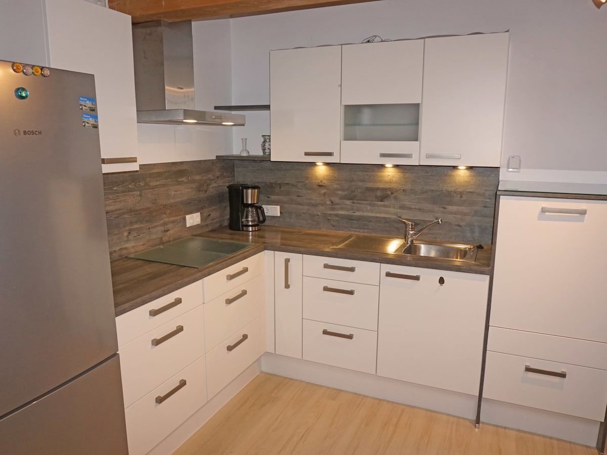 A modern kitchen features sleek white cabinetry and a wooden countertop. Stainless steel appliances, including a refrigerator and a built-in oven, are visible. Under-cabinet lighting illuminates the workspace, enhancing visibility. A coffee maker is placed on the counter, contributing to the functional design.