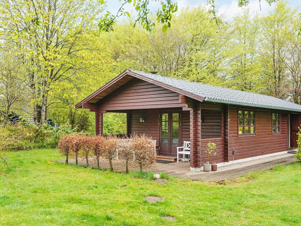 A wooden holiday home is surrounded by lush greenery and features a spacious terrace in front. Large windows allow natural light to illuminate the interior. A neatly trimmed hedge borders the property, enhancing the tranquil outdoor setting.