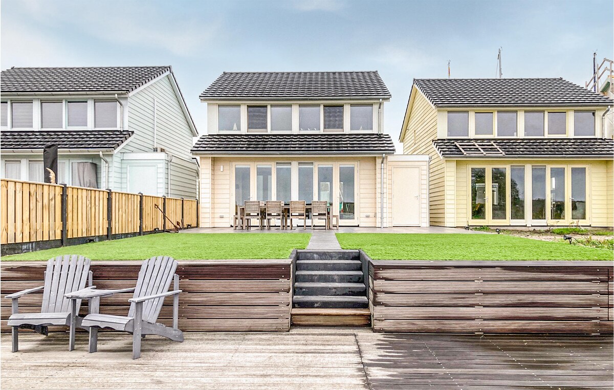 A spacious villa features two stories with multiple large windows allowing natural light to fill the interior. The patio area is adorned with wooden chairs and a green lawn, leading down to a wooden deck that provides a relaxing outdoor space.