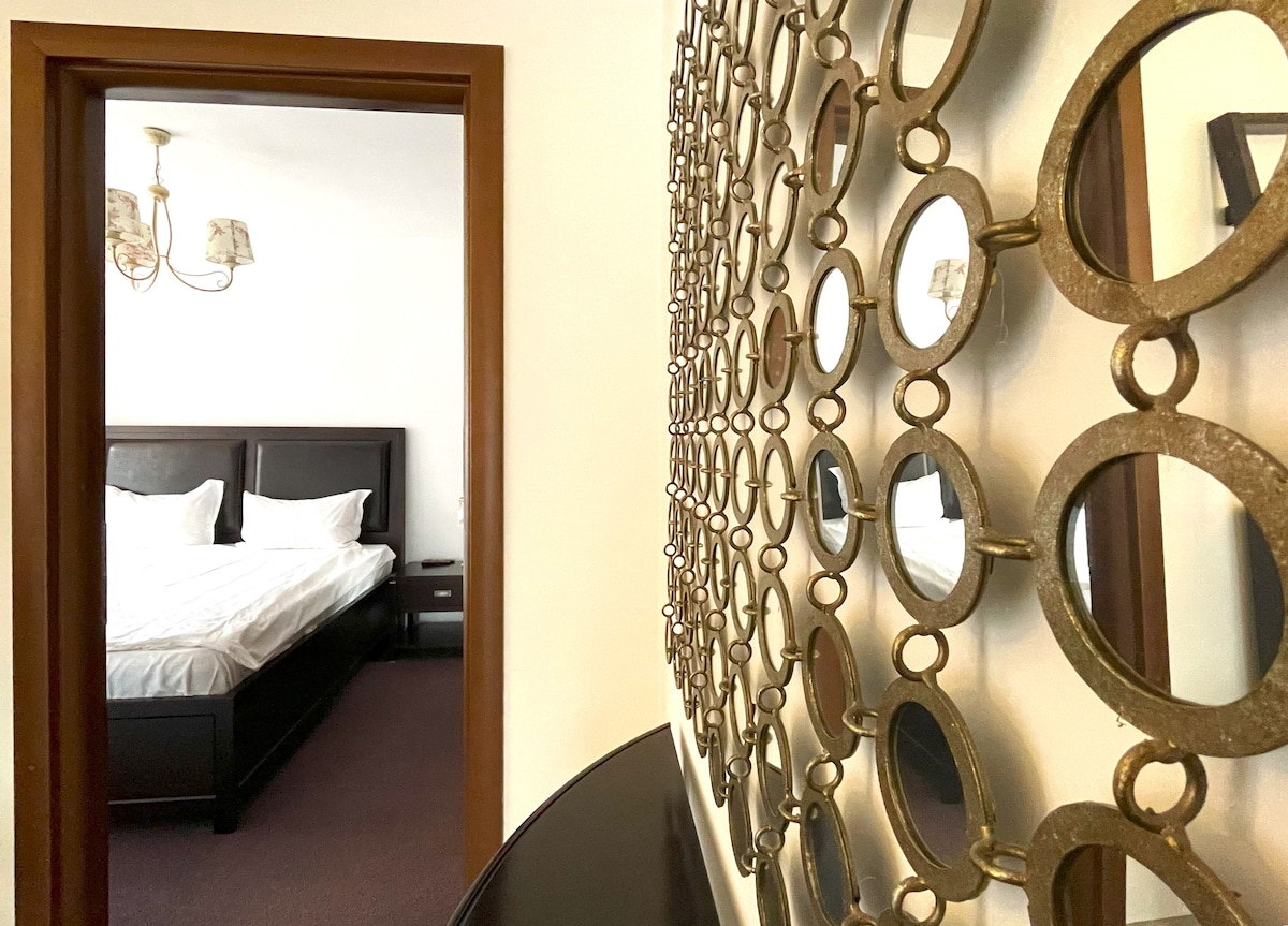 A hallway leading to a bedroom is framed by a decorative mirror featuring circular elements. The spacious bed is visible through the doorway, complemented by bedside tables. Soft lighting from a chandelier casts a gentle glow, enhancing the inviting atmosphere.