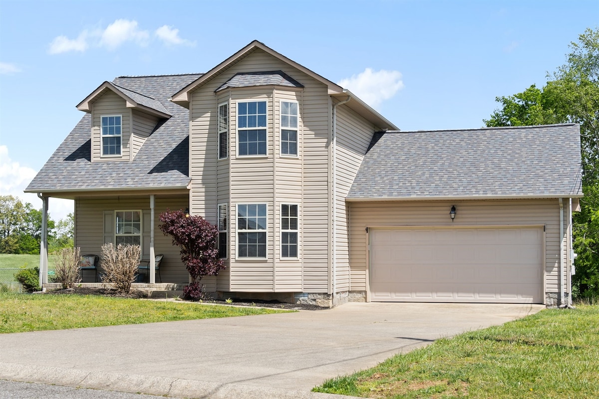 The exterior of the home is showcased, featuring a two-story structure with light-colored siding and a pitched roof. Large windows provide ample natural light, while a neatly maintained lawn and driveway add to the curb appeal. A garage with a closed door is visible on one side.