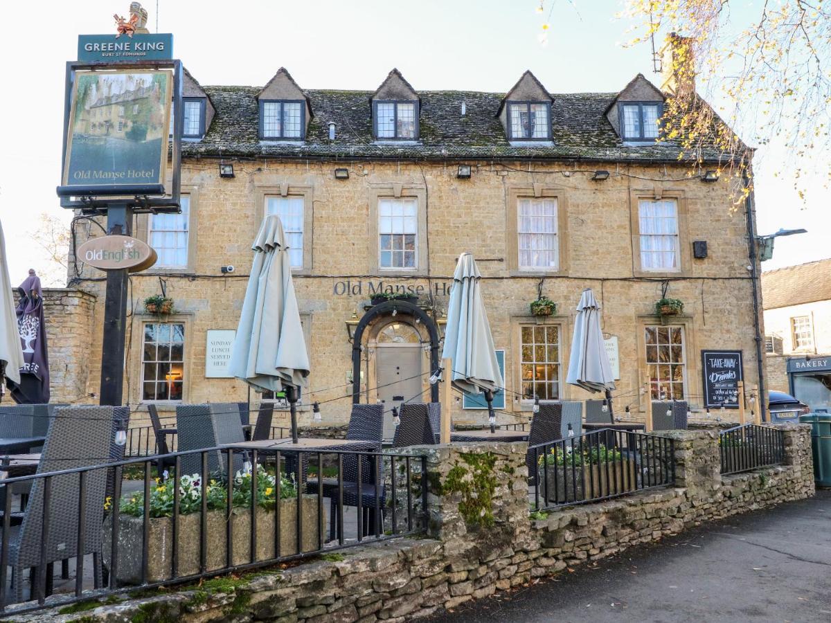 The exterior of a historic building is shown, featuring stone walls and a slate roof. The entrance is adorned with planters and outdoor seating, complete with umbrellas. Signage indicates it is the Old Manse Hotel, suggesting a welcoming atmosphere for visitors.