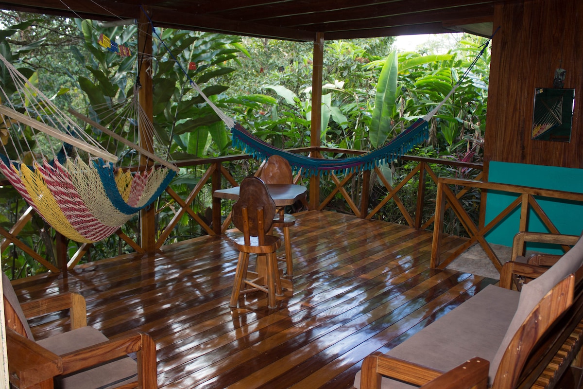 A wooden terrace is seen, featuring a polished floor and surrounded by lush greenery. Two colorful hammocks are suspended between wooden posts, providing a cozy resting area. A bamboo table and wooden chairs are positioned centrally, inviting relaxation and enjoyment of the natural surroundings.