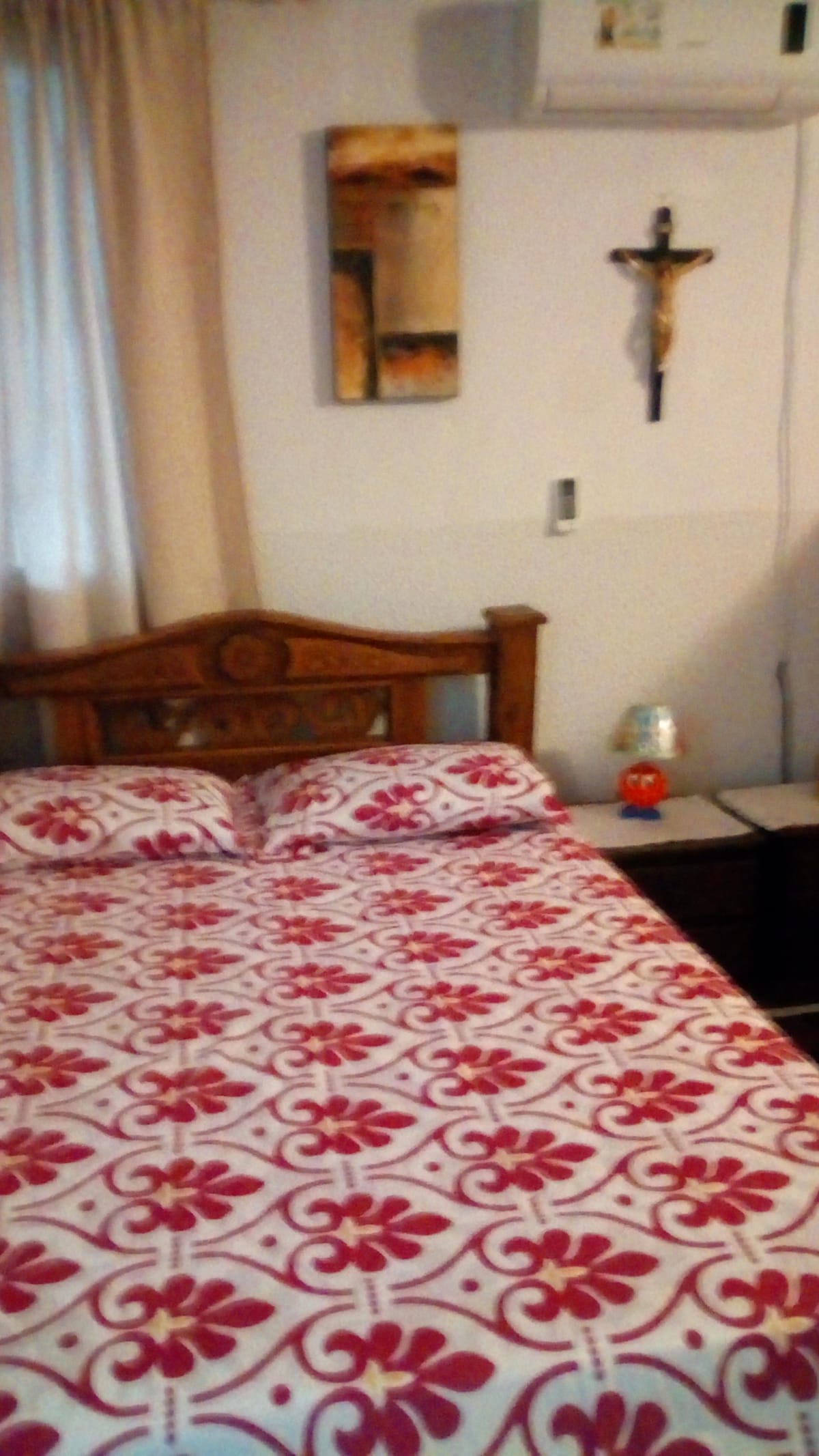 A bed with a patterned red and white bedspread is positioned against a wall. Flanking the bed are two nightstands. An air conditioning unit is mounted above, and a piece of artwork along with a crucifix decorates the wall. Natural light filters through sheer curtains.