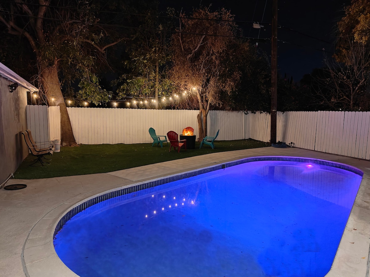The heated pool is illuminated in a calming blue hue, surrounded by a concrete patio. Nearby, an outdoor firepit is framed by colorful chairs under string lights, enhancing the inviting atmosphere of the backyard.