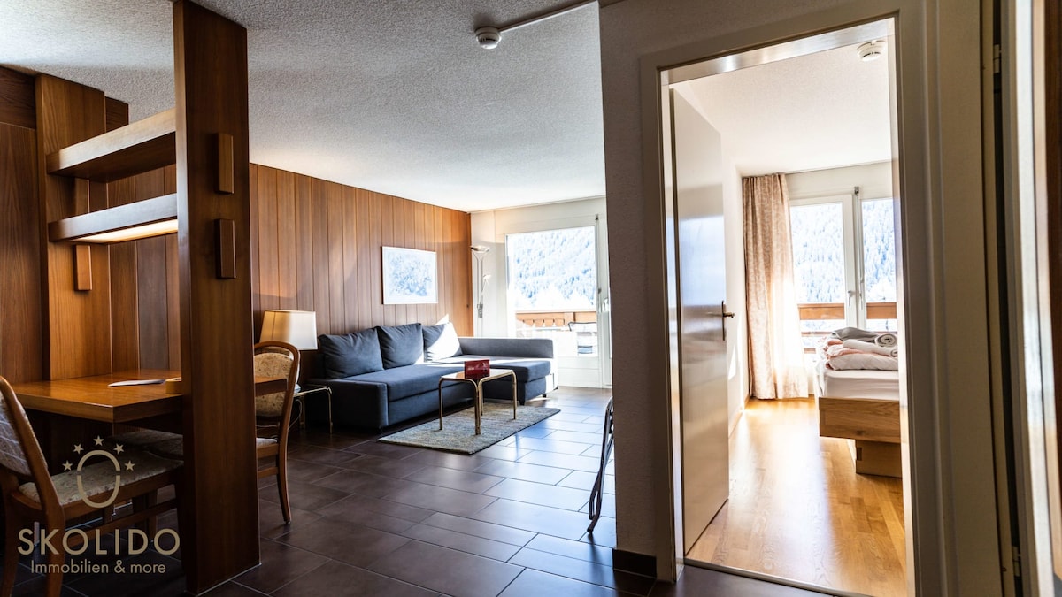 A spacious living area is visible, featuring a dark sofa and a light area rug. A dining table with two chairs is positioned near wooden paneling. Natural light pours in through large windows, illuminating a doorway leading to a separate sleeping area with a cozy bed.