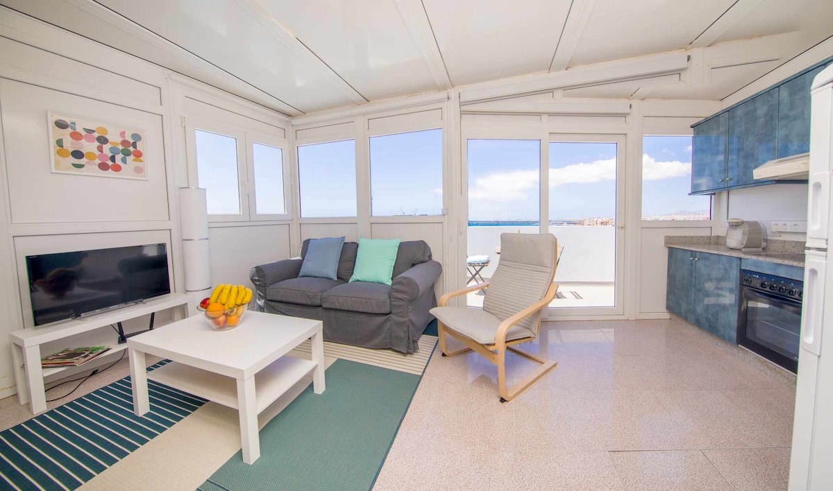 A cozy living area is illuminated by natural light streaming through large windows, offering views of the sea. A comfortable sofa is paired with a relaxing chair and a coffee table, which holds a bowl of fresh fruit. A fully equipped kitchen is visible in the background.
