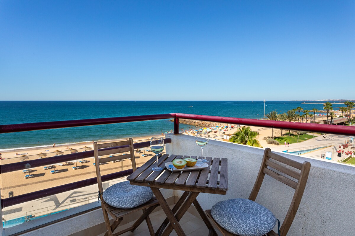 A small wooden table with two circular cushions is seen on a balcony overlooking a sandy beach and the ocean. Glasses of white wine are placed on the table alongside a plate of food, while vibrant blue skies contrast with the sea.