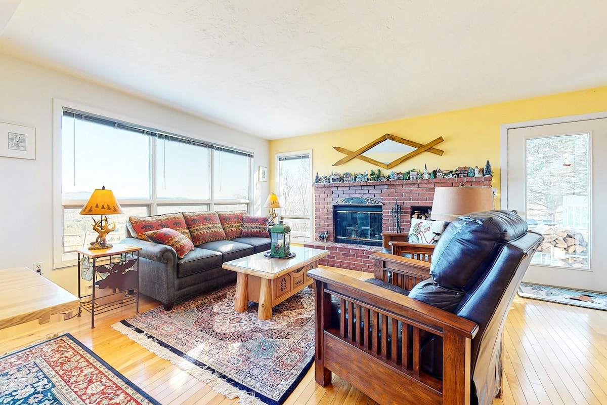 A bright living area features a comfortable sectional sofa adorned with colorful cushions, a wooden coffee table, and a brick fireplace. Large windows offer views of the outdoors, while a wooden side table holds a lamp. A cozy rug adds warmth to the hardwood floor.