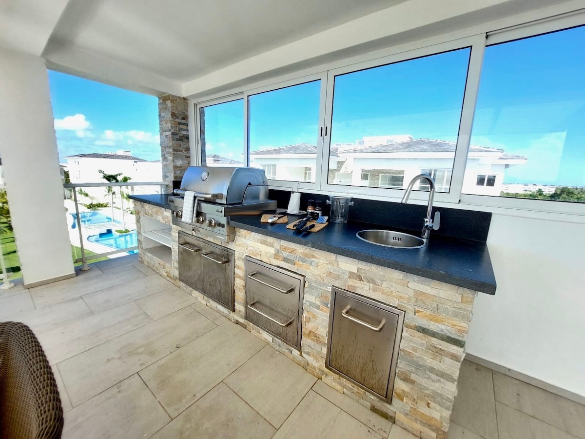 An outdoor kitchen area features modern appliances including a grill and a sink, complemented by a stone accent wall. Large windows provide a view of the surrounding landscape, ensuring a bright and airy space for cooking and entertaining.