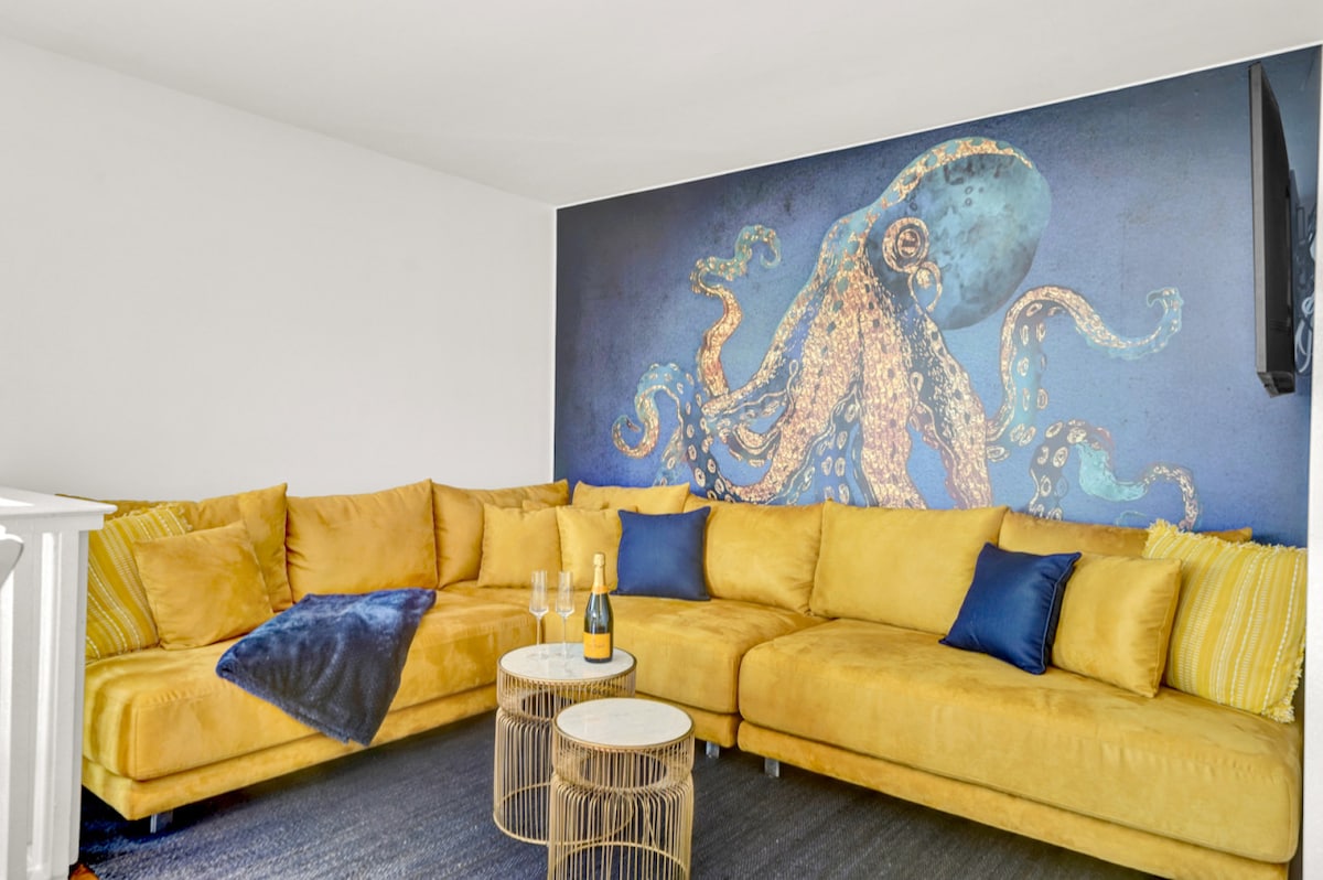 A spacious living area features a large yellow sofa adorned with blue accent pillows. A vibrant octopus mural decorates the wall, complemented by modern coffee tables and a flat-screen TV. Natural light enhances the inviting atmosphere of the room.