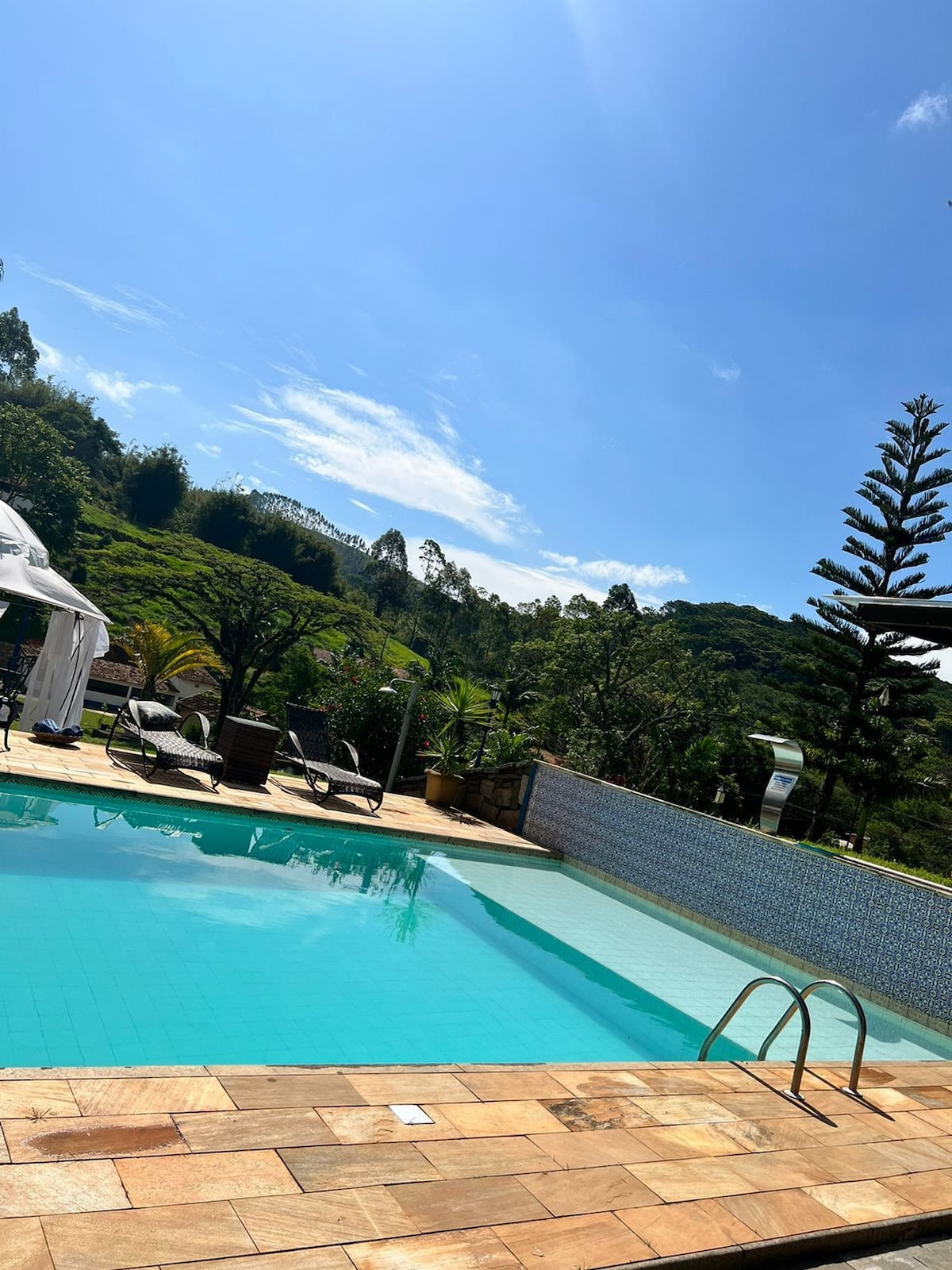 A calm pool area features a clear blue pool surrounded by stone decking. Lounge chairs are positioned beside the water, with a canopy providing shade. Lush greenery and trees are visible in the background against a bright blue sky.