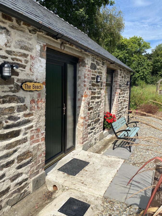 A rustic stone exterior is depicted, featuring a welcoming entrance with a small sign reading 'The Beach.' Two green chairs are placed outside, alongside vibrant red flowers in bloom. Natural greenery surrounds the structure, creating a serene setting.