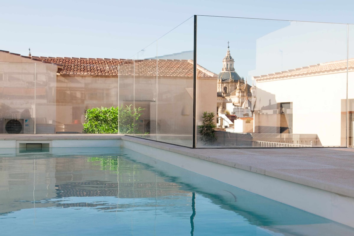 A rooftop pool is visible, surrounded by glass partitions, providing an unobstructed view of the surrounding cityscape. The reflection of a nearby church dome can be seen on the water's surface, with clear blue skies above and terracotta rooftops in the background.