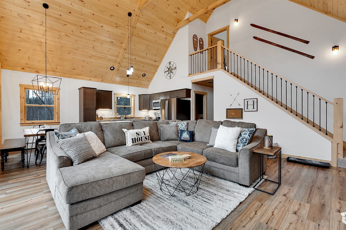 The spacious living area features a large, comfortable sectional sofa adorned with decorative pillows. Light wood paneling enhances the warm atmosphere, while a round coffee table rests on a soft area rug. A dining space and a well-equipped kitchen are visible in the background.