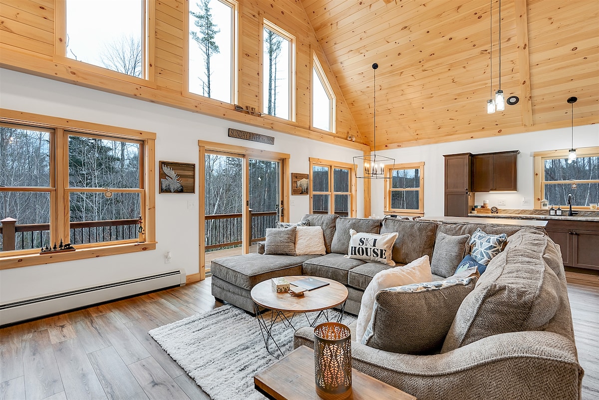 The open living area features a spacious sectional sofa arranged around a wooden coffee table, accented by a textured rug. Large windows allow natural light to fill the room, showcasing views of the surrounding trees. The kitchen is visible in the background, offering modern cabinetry.