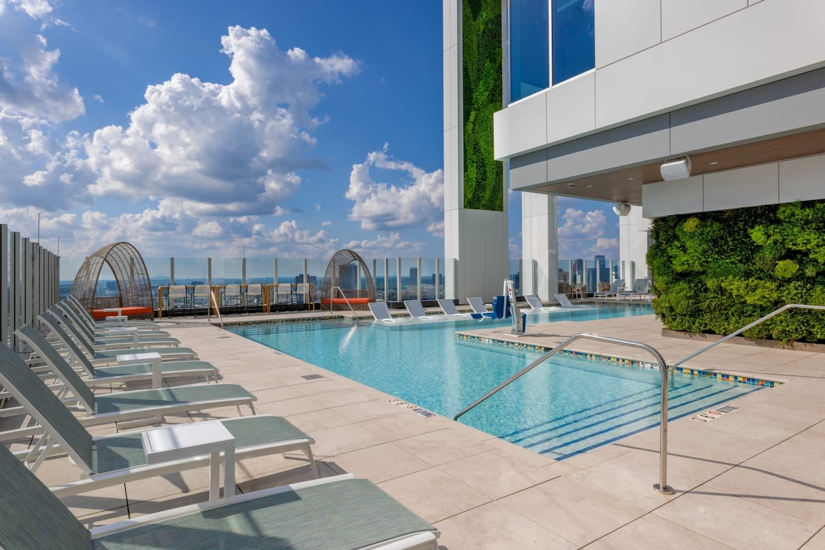 An outdoor swimming pool is surrounded by lounge chairs, with a modern building and green wall features in the background. The sky is bright with scattered clouds, enhancing the inviting atmosphere for relaxation and leisure.