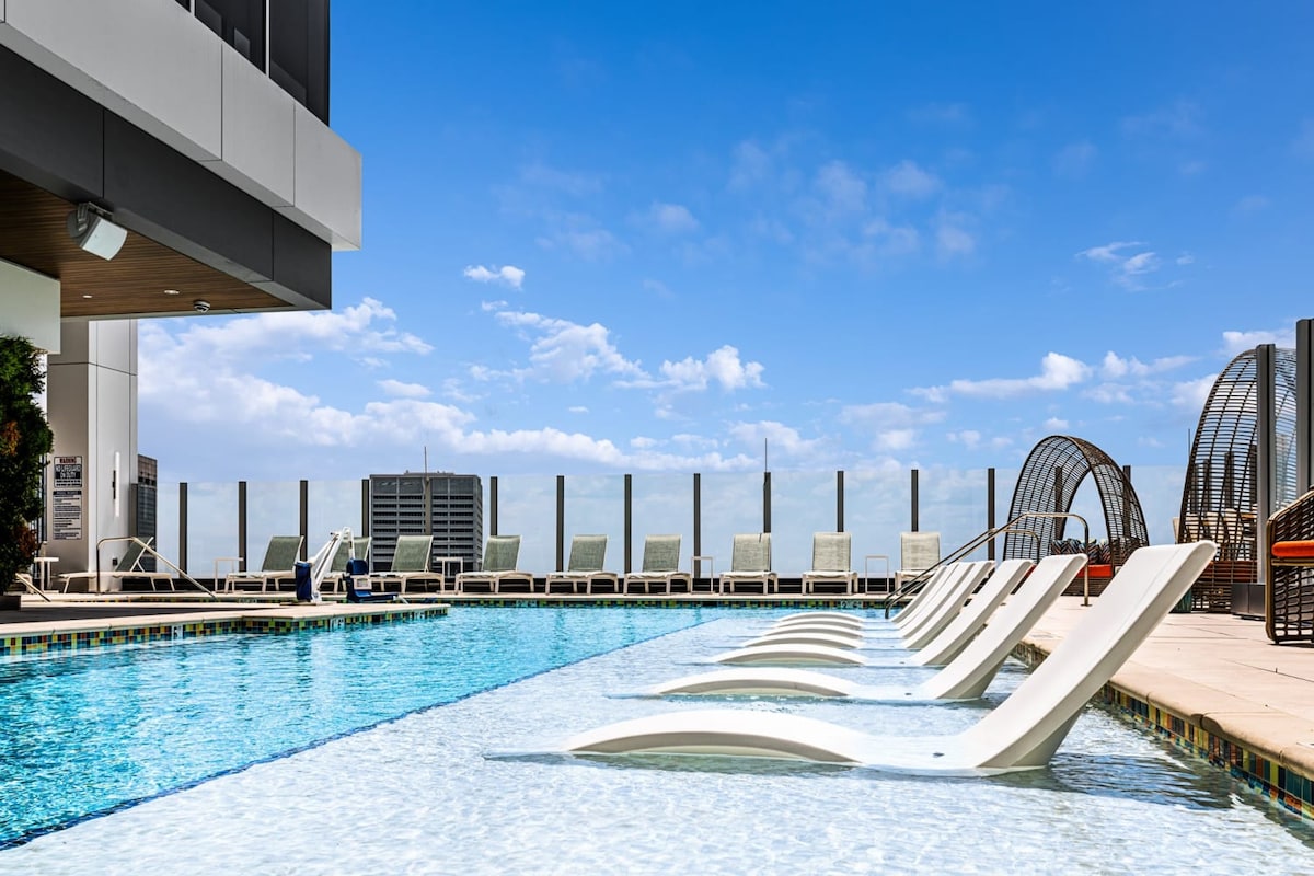 An outdoor swimming pool is set against a clear blue sky, featuring sleek white lounge chairs positioned along the water's edge. The area is surrounded by a modern railing, with additional seating visible in the background, inviting relaxation and leisure.