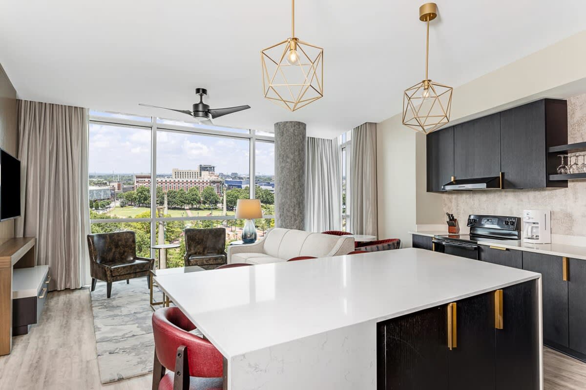 A bright and airy living space features large windows offering a view of the surrounding landscape. Modern furnishings include a spacious white sofa and dark wood chairs. The kitchen area showcases a white countertop and sleek cabinetry, complemented by decorative light fixtures hanging overhead.