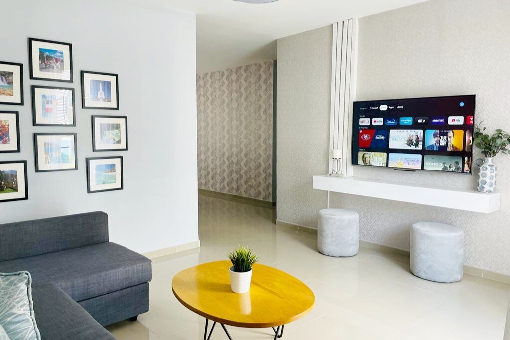 A modern living area features a gray sectional sofa and a round yellow coffee table. A wall-mounted TV displays entertainment options, while framed photographs adorn the adjacent wall. Two light gray stools add seating near the TV, enhancing the space's functionality.
