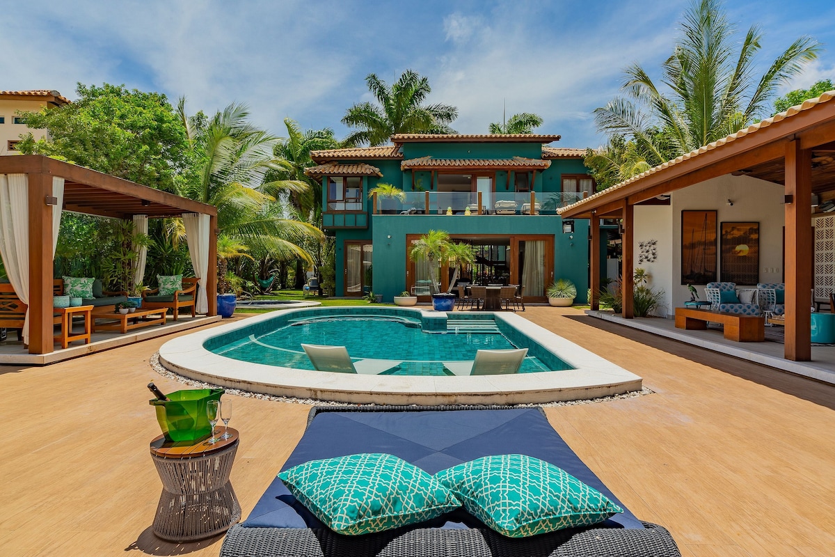 An inviting outdoor space is captured, featuring a private pool surrounded by tropical greenery. Cozy lounge chairs are positioned by the water, with shaded seating areas visible in the background. The vibrant colors of the house complement the lush landscape, creating a serene atmosphere.