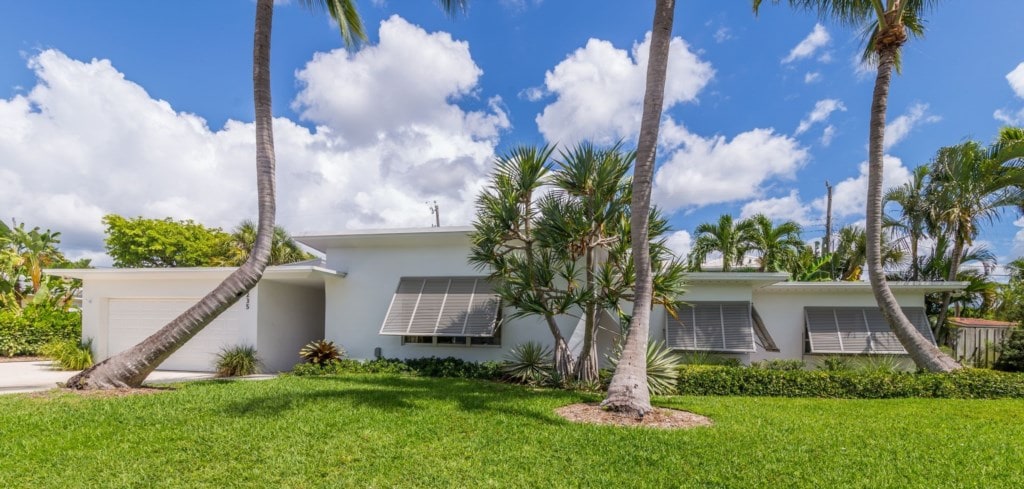 A mid-century modern home is framed by lush green landscaping and palm trees. Large windows are fitted with awnings, allowing natural light while providing shade. A driveway leads to a two-car garage, with a well-maintained lawn enhancing the inviting exterior.