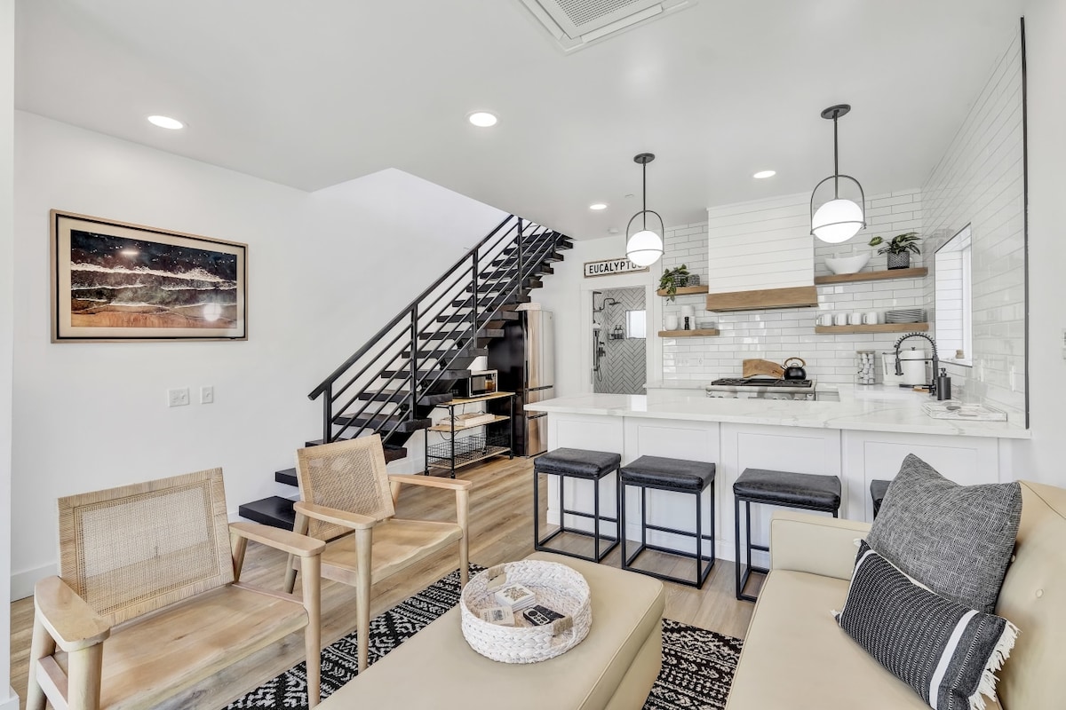 A modern living area contains a comfortable seating arrangement with neutral tones. An open kitchen features a white tile backsplash and bar seating, while a staircase with sleek black railings provides visual interest. Natural light fills the space, highlighting the design elements.