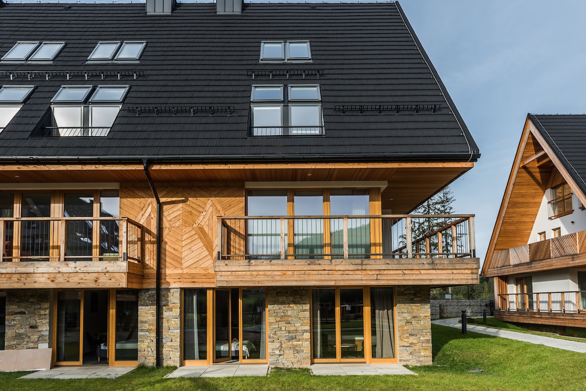 A modern multi-level villa showcases a wooden facade with stone accents, featuring large glass doors and expansive balconies. A manicured green lawn surrounds the building, providing an inviting outdoor space. The slanted roof design complements the mountain atmosphere of the surrounding area.