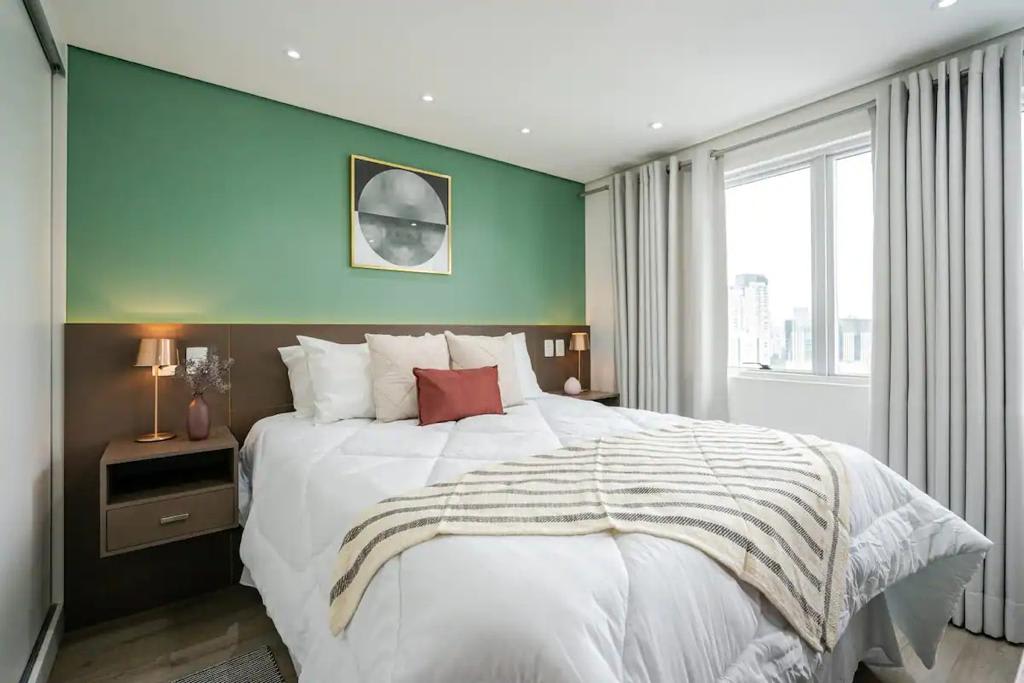 Ელეგანტური ოთახი ორადგილიანი საწოლით, ყავისფერი თავსაფრით, მწვანე კედლით და მინიმალისტური დეკორით. Საწოლს აქვს თეთრი საწოლის გადასაფარებელი, თეთრი ბალიშები და წითელი. Მეზობლად, თანამედროვე ლამპები და დიდი ფანჯარა ნაცრისფერი ფარდებით, რომელიც უზრუნველყოფს ბუნებრივ განათებას.