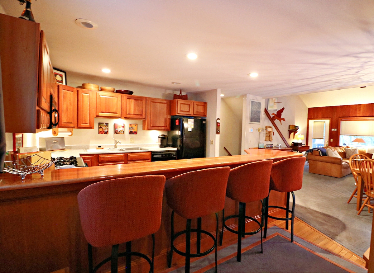 A warm kitchen area is highlighted by wooden cabinetry and a spacious countertop. Four bar stools are arranged at the breakfast bar, providing casual seating. The kitchen is equipped with modern appliances and soft lighting that enhances the inviting space.
