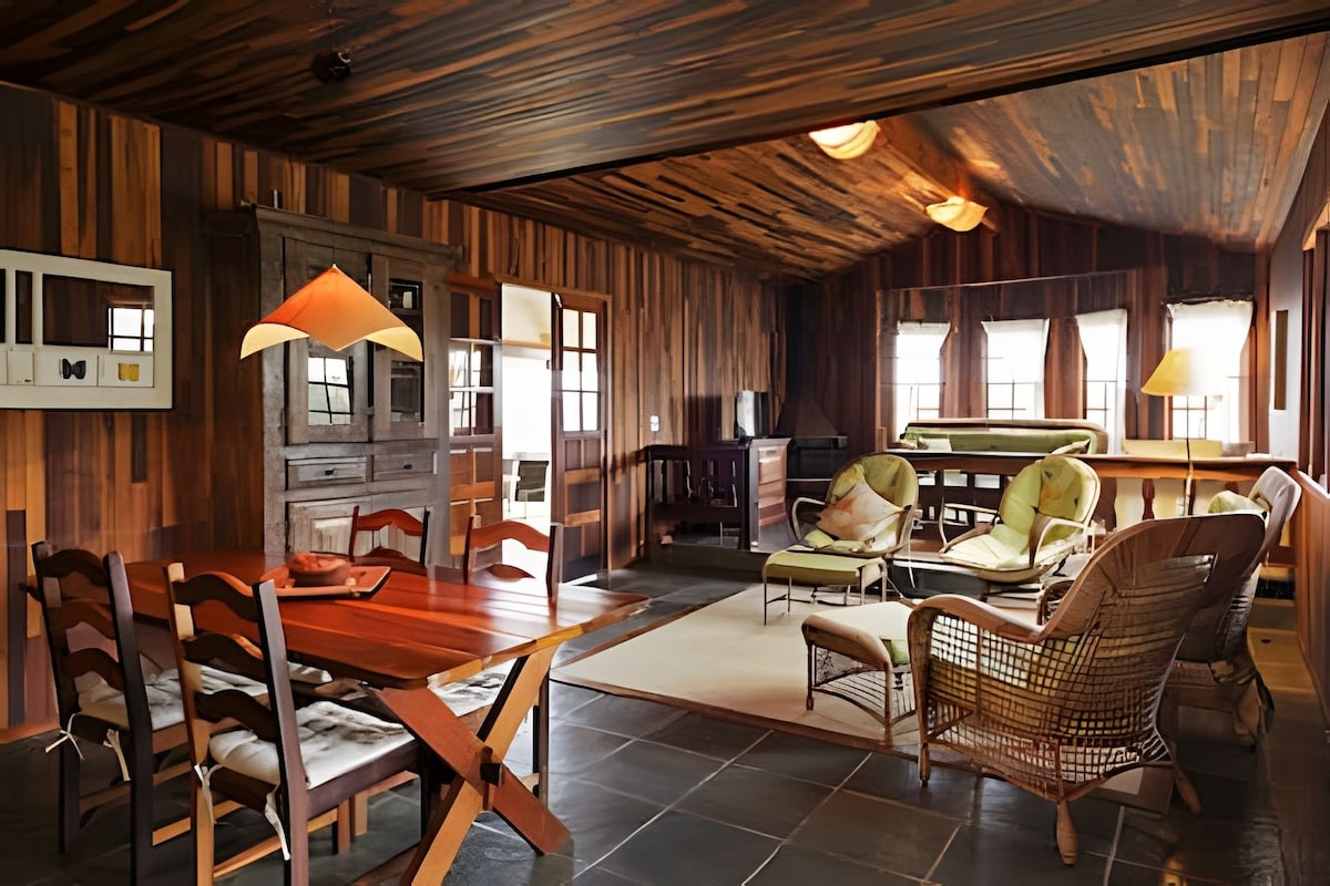 The interior features warm wooden walls and a ceiling, creating a cozy ambiance. A wooden dining table with chairs is positioned to one side, while a seating area with comfortable armchairs and a sofa is visible. Large windows allow natural light to fill the space.