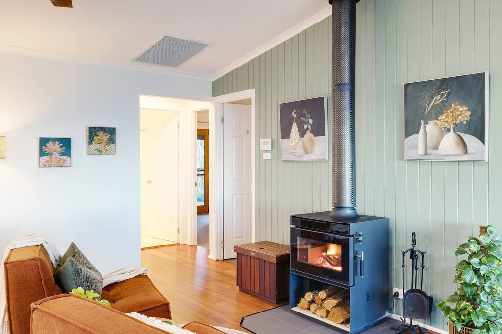 A cozy living area features a wood-burning fireplace framed by green panel walls. The space is enhanced by warm wood flooring and modern artwork, creating an inviting atmosphere. A comfortable sofa is positioned nearby, inviting relaxation on cooler evenings.