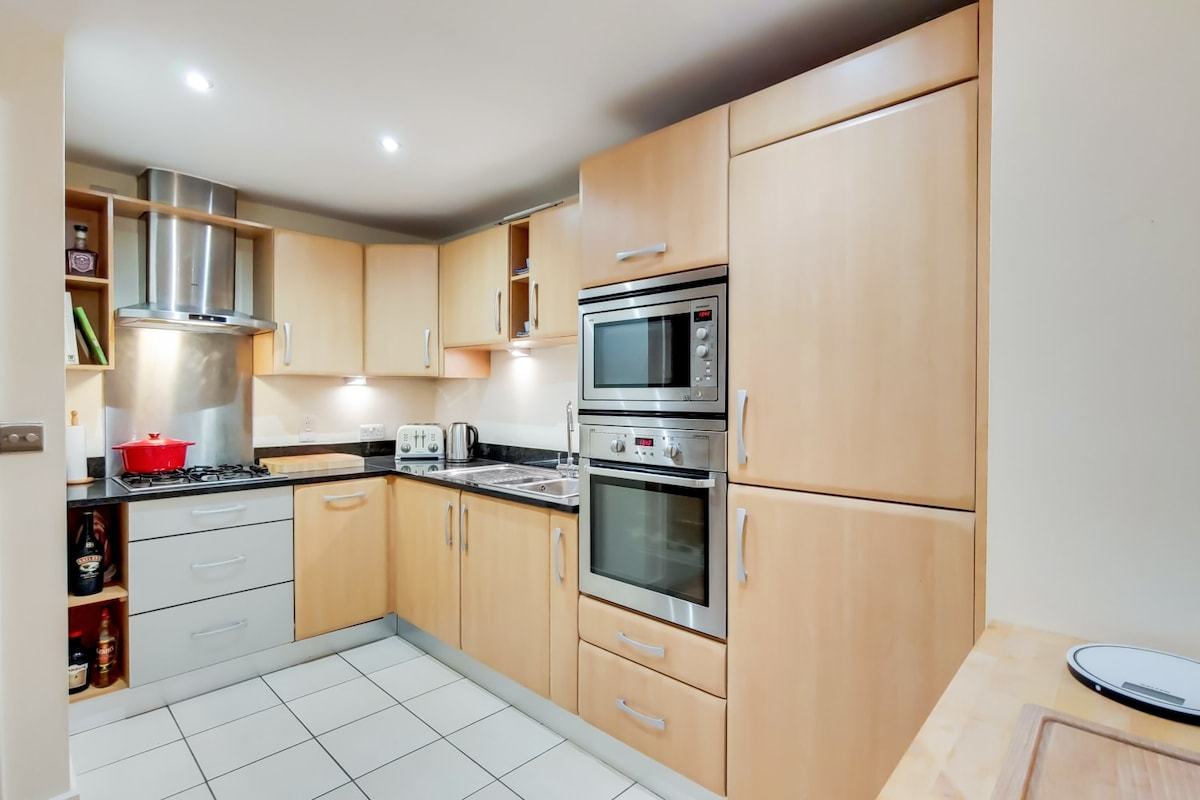 A modern kitchen is presented with wood cabinetry and sleek black countertops. Stainless steel appliances, including a gas stove and double wall oven, add functionality. Under-cabinet lighting highlights the cooking space, while tiled flooring offers easy maintenance throughout.