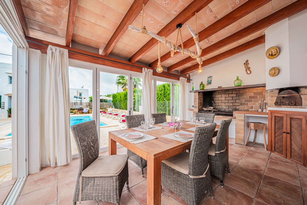 The dining area is furnished with a wooden table surrounded by six wicker chairs. Large glass doors allow natural light to fill the space, offering a view of the outdoor pool area. Soft white curtains frame the windows, and a stone barbecue is visible nearby.
