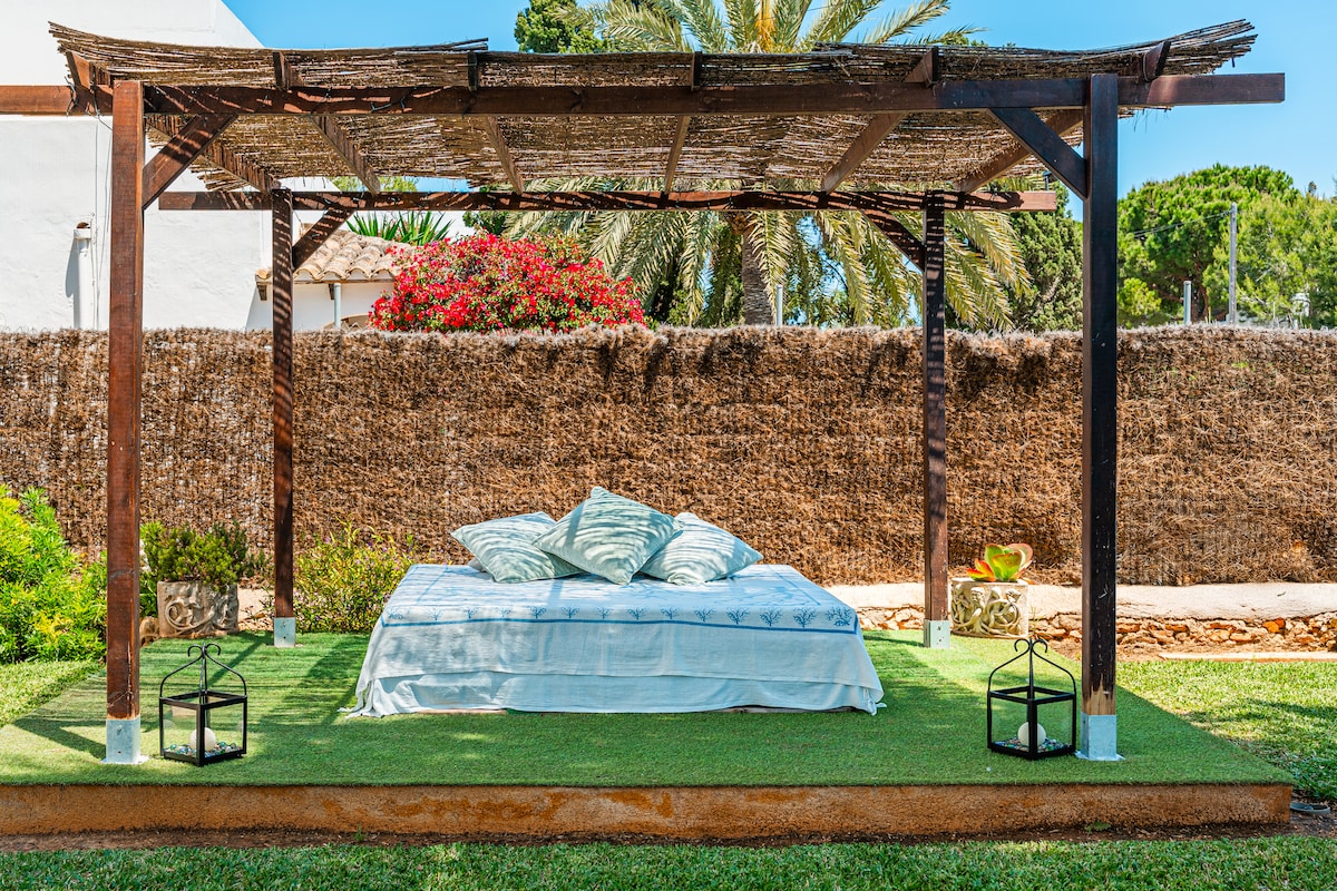 A shaded outdoor area features a comfortable bed adorned with decorative cushions, set on a patch of green turf. Lanterns are positioned on either side, providing a serene and inviting spot for relaxation amid lush vegetation and a backdrop of a natural fence.
