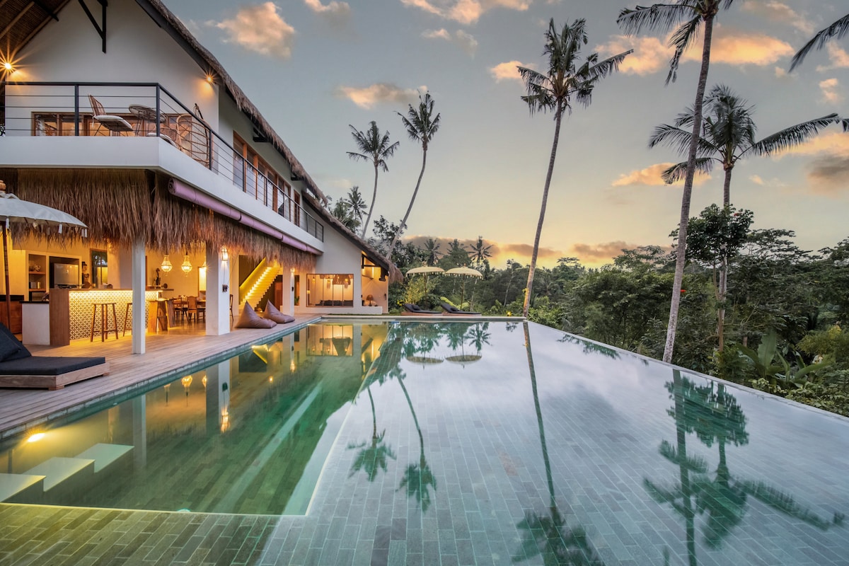 The villa's infinity pool reflects the serene jungle surroundings, with tall palm trees framing the view. An open-air living area is visible, designed to blend seamlessly with nature, while the warm evening light enhances the inviting atmosphere.
