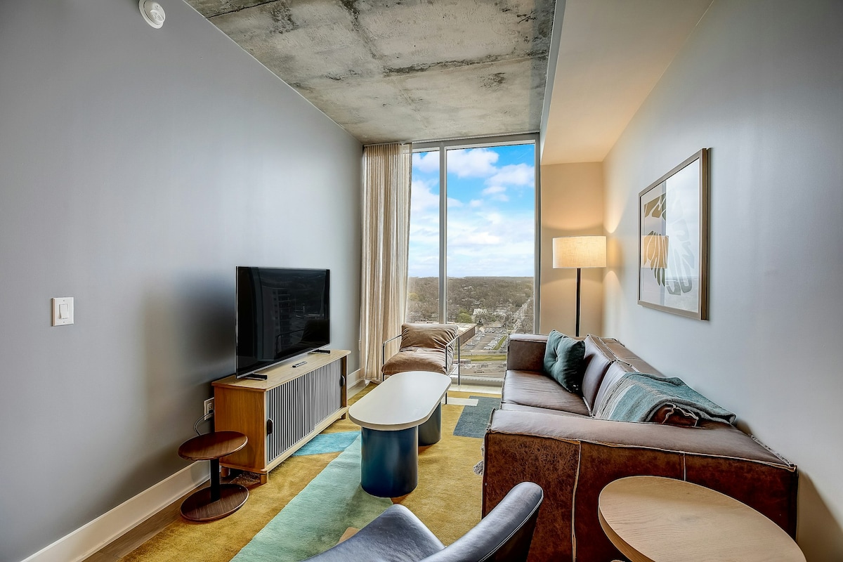 A modern living area features large floor-to-ceiling windows that let in ample natural light. A plush sofa and accent chair are arranged around a minimalist coffee table. A sleek TV console sits opposite the seating area, enhancing the contemporary design.