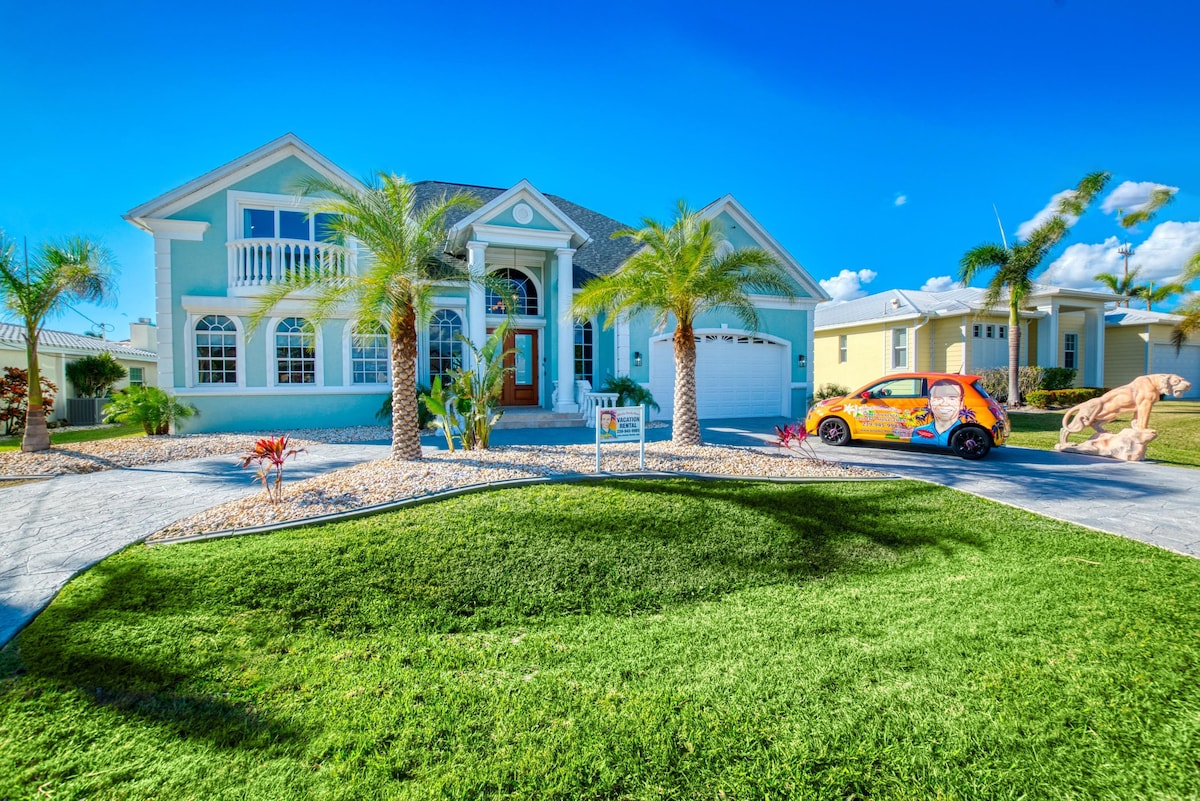 A large light blue home is framed by neatly manicured lawns and colorful landscaping. Palm trees line the driveway, leading to a welcoming entrance with an arched doorway. A small, vibrant car is parked in the driveway, adding a cheerful touch to the outdoor scene.