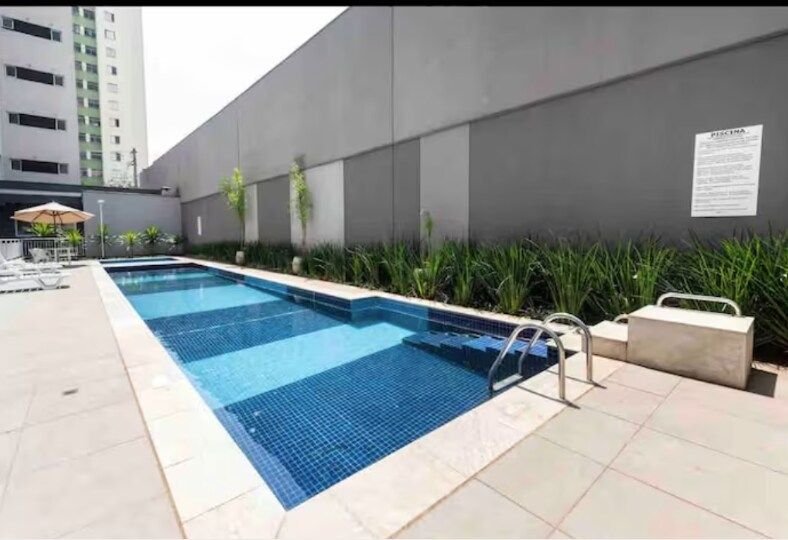 A long, narrow pool is framed by tiled areas and lush green landscaping. Grey walls provide a modern backdrop, while lounge chairs and an umbrella are positioned nearby for relaxation. The water is a clear blue, reflecting the surrounding light.