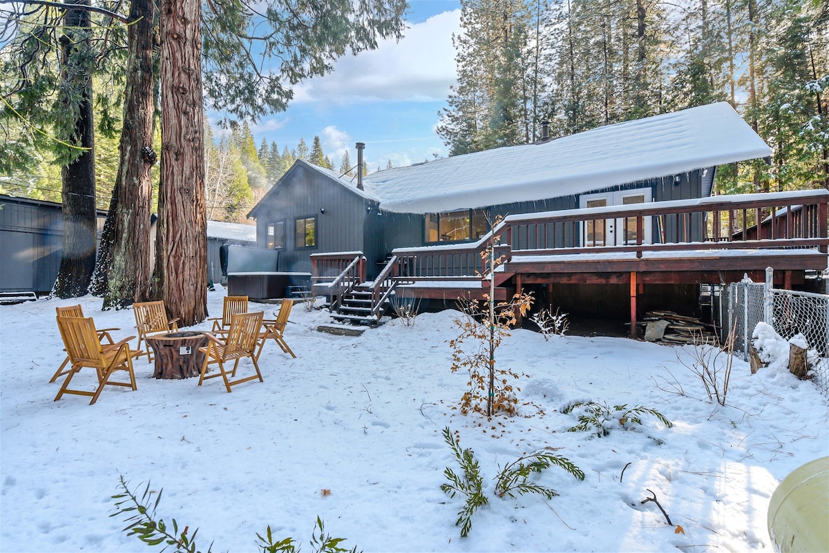 A cozy cabin is surrounded by a snowy landscape, featuring a wooden deck that extends from the entrance. Two Adirondack chairs are situated around a fire pit in the backyard. Towering evergreen trees provide a natural backdrop, enhancing the serene setting.