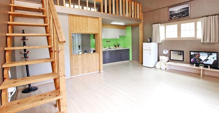 A spacious and open living area features wooden accents and high ceilings, with a staircase leading to a lofted sleeping area. A functional kitchen with modern appliances is visible, along with a refrigerator. Natural light streams through large windows, illuminating the floor.