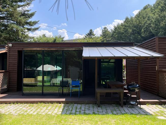 An outdoor view of a wooden property featuring large glass doors that open to a spacious deck. Seating arrangements include a table with several chairs, and a covered area is provided for shaded relaxation. A barbecue grill is positioned nearby, surrounded by lush greenery.