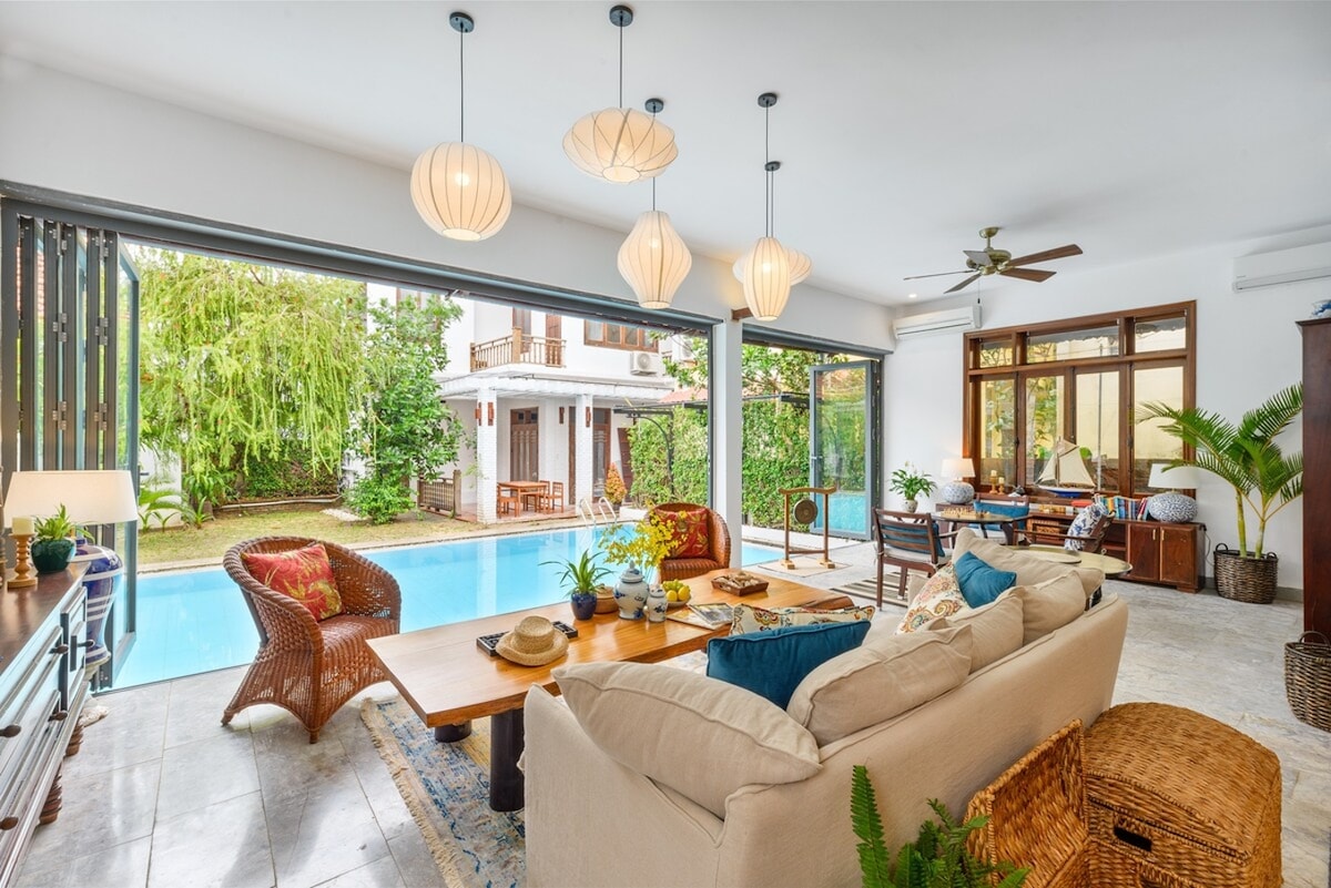A spacious living area features large sliding glass doors opening to a lush garden and pool. A comfortable sofa is positioned near a wooden dining table, adorned with decorative items and plants. Light fixtures hang from the ceiling, providing a warm ambiance.