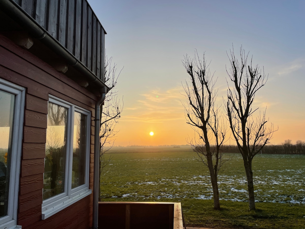 The image captures a sunset view from the accommodation, featuring a warm orange glow on the horizon. Two bare trees are visible in the foreground, with expansive green fields stretching out into the distance. Large windows of the building reflect the evening light.