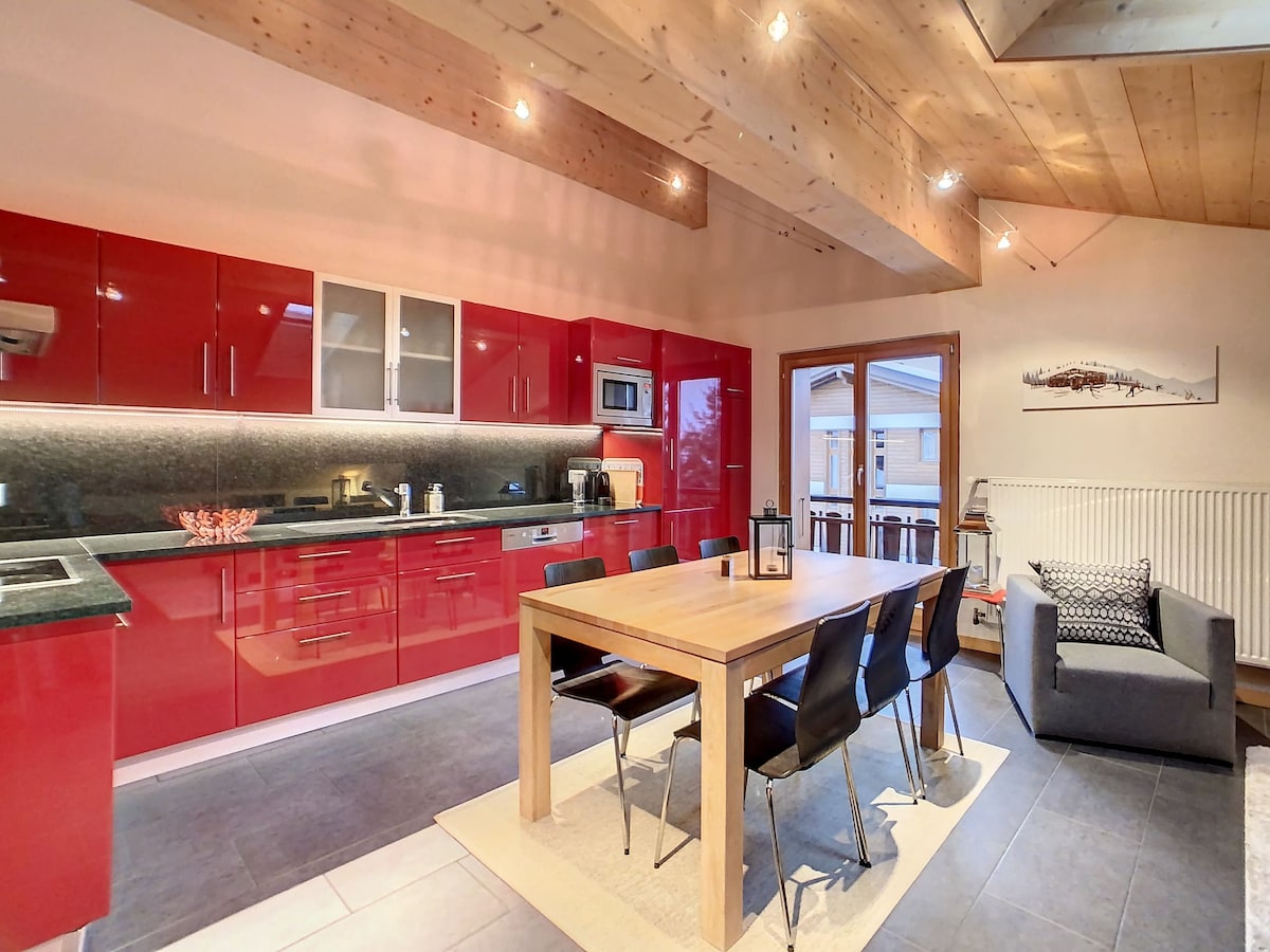 The kitchen and dining area feature modern red cabinetry and a spacious wooden dining table, accommodating multiple chairs. A cozy seating area is visible, with a grey armchair positioned nearby. Natural light fills the space through a large window, enhancing the inviting ambiance.