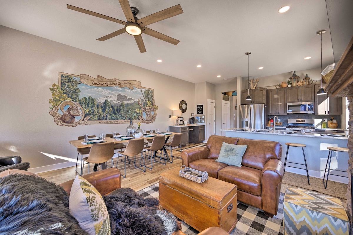 An inviting living space features a comfortable leather sofa paired with a decorative box and a textured throw. A large dining table with place settings is visible, along with a mural showcasing mountain scenery. A modern kitchen with stainless steel appliances can be seen in the background.